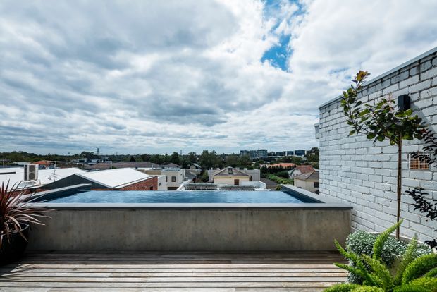 Rooftop terrace with a narrow rectangular swimming pool, timber decking, a concrete pool wall, and planted greenery. The view looks across a low-rise urban residential neighbourhood toward distant apartment buildings beneath a cloudy blue sky. A white-painted brick wall forms part of the terrace enclosure.