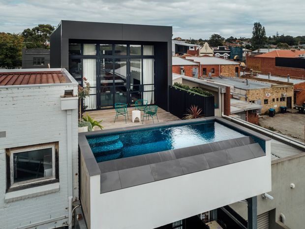 Elevated exterior view of a contemporary urban residential extension with a rooftop terrace and swimming pool. The dark-framed glazed pavilion opens onto a timber deck with outdoor seating, while the pool sits on a projecting white structure above the surrounding buildings. The dense suburban setting includes visible brick and rendered neighbouring structures under an overcast sky.