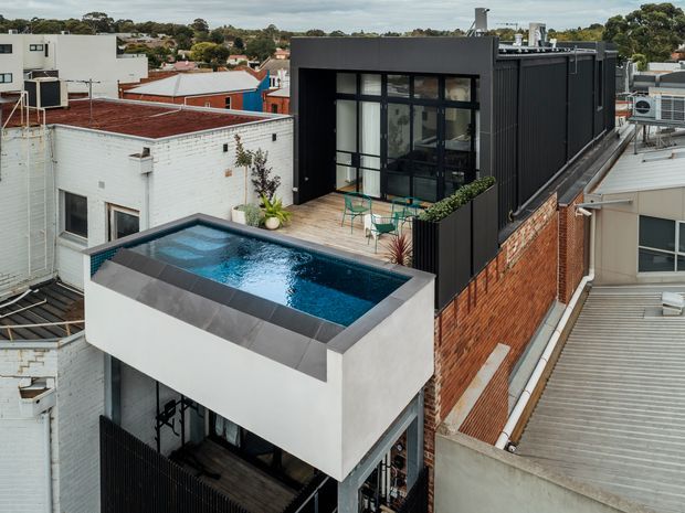 Elevated view of a compact urban rooftop addition with a raised rectangular swimming pool, dark tiled coping, and a glazed black-framed pavilion opening onto a small terrace. The extension combines white rendered walls, dark vertical cladding, exposed brickwork, and extensive glazing, surrounded by neighbouring low-rise buildings and rooftops.