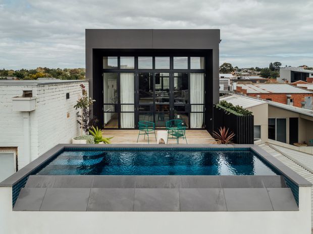 A rooftop terrace with a rectangular swimming pool in the foreground, surrounded by large grey tiles. Beyond the pool is a raised deck with outdoor chairs and a small table, opening into a dark-framed glazed pavilion or upper-level living space. The surrounding urban rooftops and cloudy sky indicate an elevated city setting. The design uses strong geometric forms, black metal-framed glazing, dark balustrades, and a restrained contemporary palette.