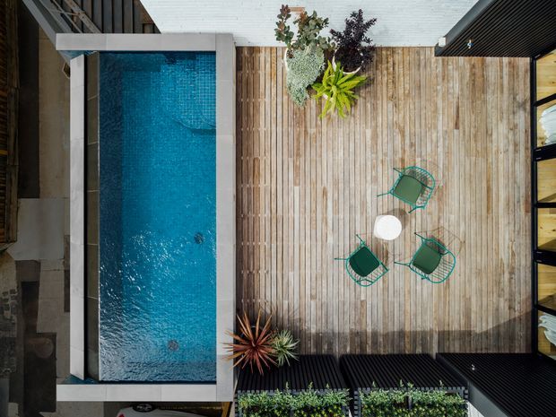Overhead view of a contemporary outdoor courtyard featuring a narrow rectangular swimming pool beside a large timber deck. The deck includes a small round table, four green wire-frame chairs, and planted pots, enclosed by dark exterior walls and vertical timber screening.
