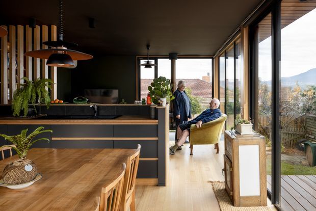 A warm, contemporary open-plan kitchen and dining area with a dark, minimalist kitchen island, timber benchtop, integrated dark cabinetry, pendant lights, and a large timber dining table. Full-height glazing along the right side opens the room visually to a timber deck and suburban landscape. A sitting area with an upholstered armchair is visible beyond the kitchen, while timber battens and indoor plants add texture and warmth.