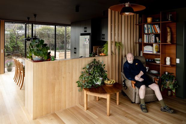 Warm, open-plan residential interior combining a kitchen and living area. Light timber flooring and a timber kitchen island lead toward full-height glazing overlooking greenery. Dark cabinetry, a vertical timber-slat feature wall, and built-in dark timber shelving create a strong contrast. The living zone includes an armchair and round timber coffee table, with abundant indoor plants and bookshelves contributing a natural, lived-in atmosphere.