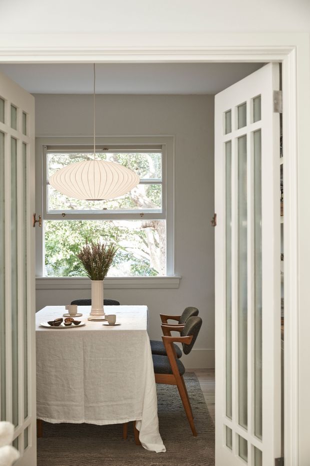 A refined dining room viewed through open glazed double doors. The room features a round or oval dining table covered with a white linen cloth, upholstered mid-century style wooden dining chairs, a large sculptural pendant light, pale grey walls, white painted trim, and a wide timber-framed window overlooking greenery. The space is softly lit and calm, with layered neutral textures and traditional architectural detailing.