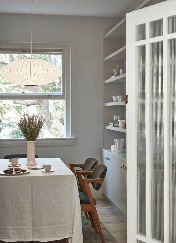 A softly lit dining area with a rectangular table covered in a light linen cloth, wooden mid-century-style dining chairs, a sculptural pendant light, and a vase of dried branches. Built-in open shelving and lower cabinetry line the wall, while a white-framed fluted-glass partition door occupies the right side. A large window brings in views of trees and natural light.