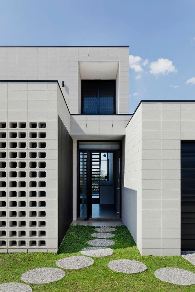 Front exterior view of a modern, rectilinear residence composed of pale modular masonry or concrete-block walls. The entry is recessed beneath projecting box forms, with dark-framed glazing and a shaded upper-level opening. A perforated breeze-block screen forms the left side of the facade, while a dark garage door is visible on the right. Circular concrete stepping stones cross a small lawn toward the entry. The composition is geometric and minimalist, photographed in bright daylight against a blue sky.