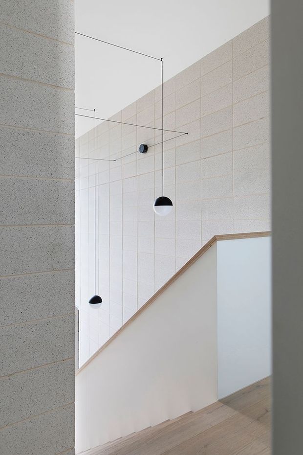Minimalist stairway or landing viewed alongside a tall, light-toned masonry wall. The space features pale timber flooring, white plaster stair walls with timber edging, exposed light concrete blockwork, and several small black-and-white pendant lights suspended on thin wires at varying heights. The composition emphasizes a bright, quiet, double-height vertical volume and geometric lines.