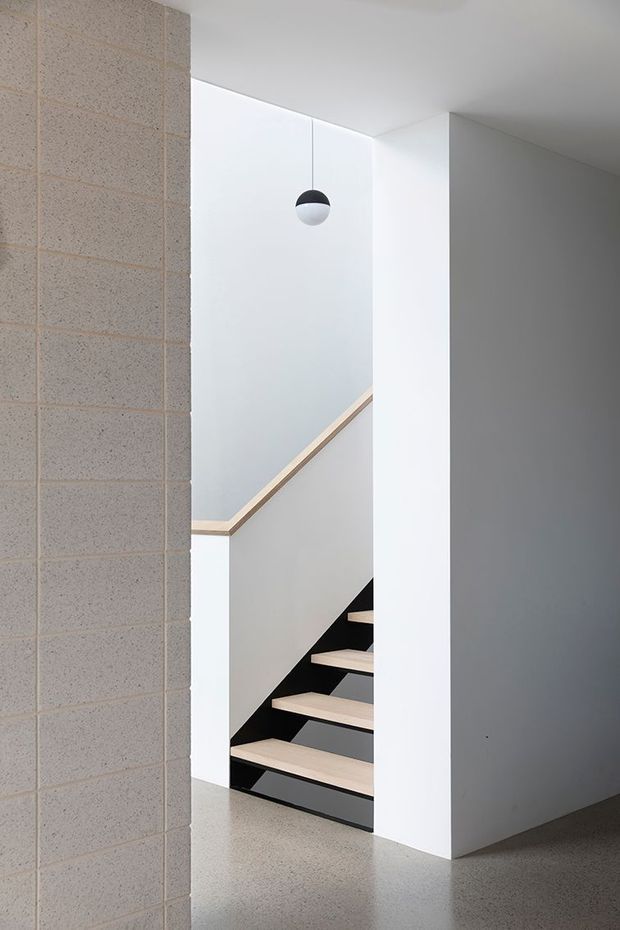 Minimalist interior view of a stairway with pale timber open-tread stairs, black steel supports, white plaster walls, and a suspended two-tone pendant light. A full-height exposed concrete block wall frames the left side, while a polished grey concrete or terrazzo-like floor continues through the space. The stairwell is bright, clean, and architecturally simple.