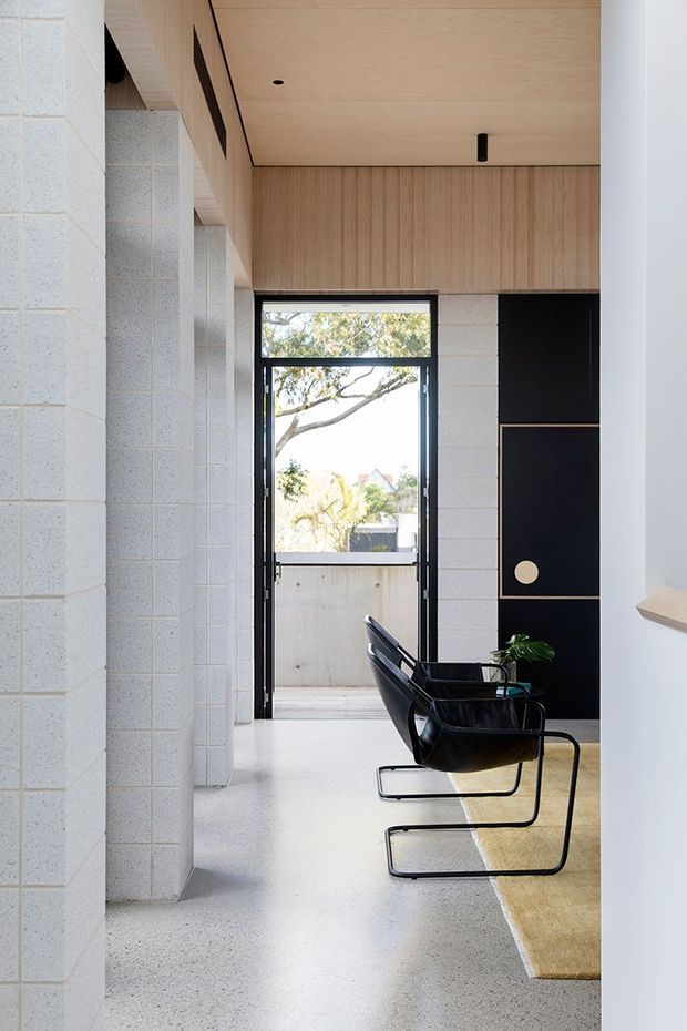 A bright, narrow modern entry hall or circulation space with exposed pale concrete block walls, a polished light terrazzo or concrete floor, and a light timber-lined ceiling and upper wall. Two black leather sling chairs with tubular metal frames sit beside a tan runner rug. A black built-in cabinet with brass or timber trim and a circular pull forms a feature wall near the glazed rear doorway. Large glazing brings in views of trees and daylight.