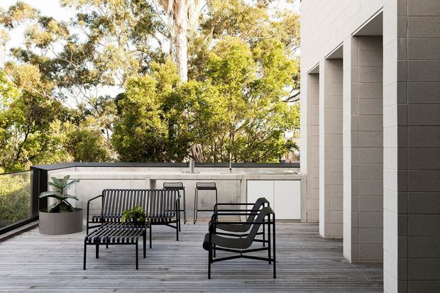 A bright contemporary outdoor terrace with grey timber decking, black metal lounge furniture, and a built-in outdoor kitchen or service counter along the rear wall. The terrace is bordered by a dark glass balustrade and framed by a pale tiled masonry facade with deep vertical openings. Mature trees are visible beyond the terrace.