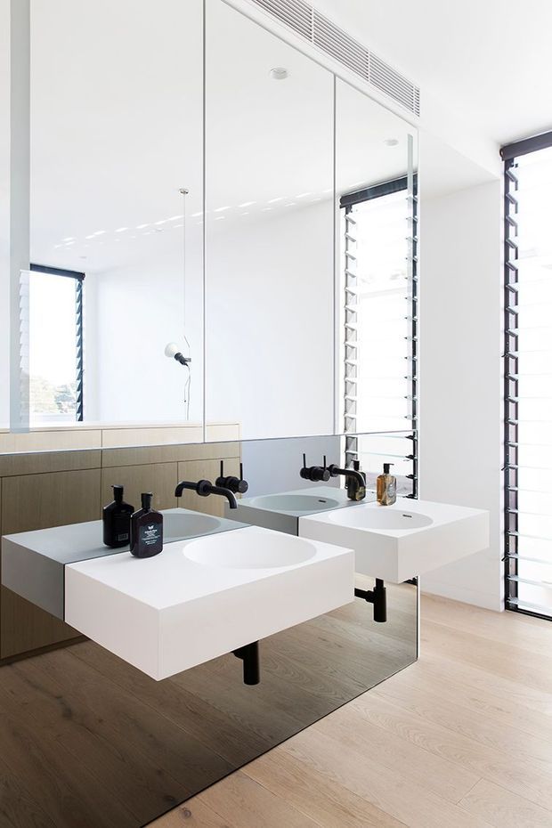 Bright contemporary bathroom with two floating white rectangular vanity basins, matte black wall-mounted taps and exposed dark plumbing. A large mirrored wall doubles the sense of space, while pale timber flooring, light wood cabinetry, white walls, and tall louvred windows create an airy minimalist interior.