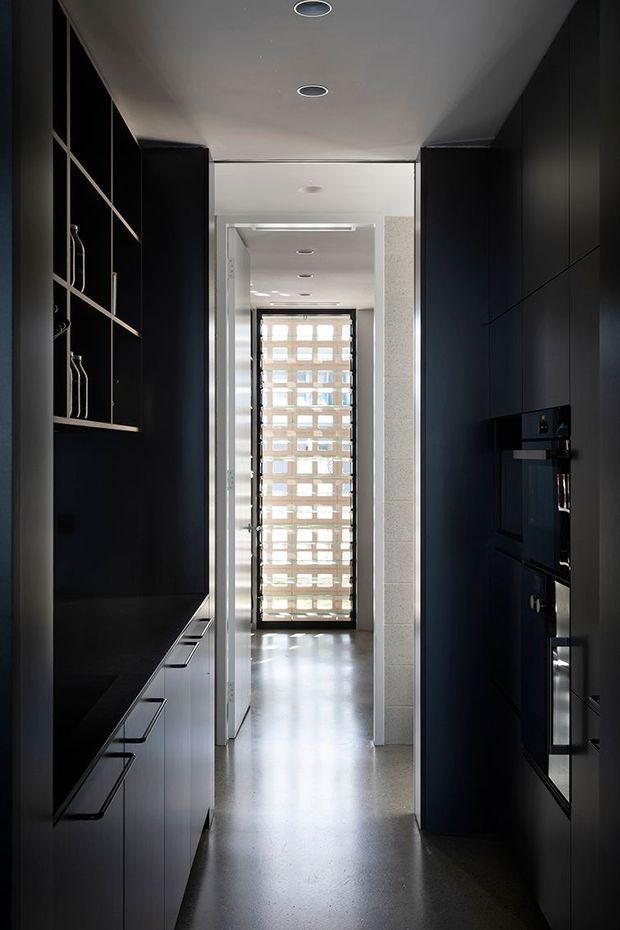 A narrow, galley-style kitchen corridor with floor-to-ceiling matte black cabinetry on both sides, integrated appliances, open shelving, dark benchtops, and slim linear handles. A bright glazed opening at the far end brings daylight into the space. The ceiling has recessed circular downlights, while the floor appears smooth and light grey, creating a high-contrast, sleek interior.