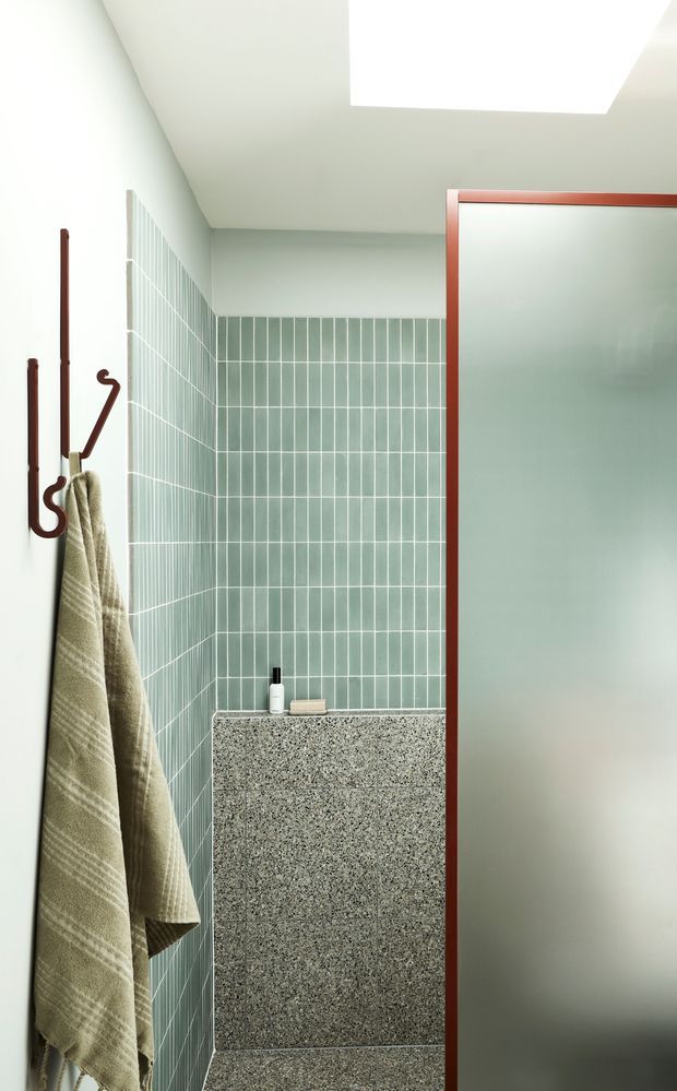 A compact, contemporary bathroom shower area with pale green vertical wall tiles, a terrazzo-clad shower ledge or half-height partition, and a frosted glass shower screen framed in dark reddish-brown metal. A skylight provides bright natural illumination, while a wall-mounted towel rail holds a neutral linen towel.