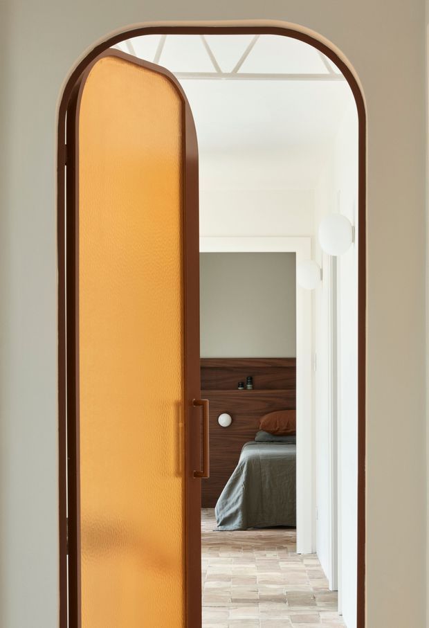 A softly lit interior view through a tall rounded-arch doorway with a warm amber translucent door and dark timber framing. Beyond the partially open door is a serene bedroom with a low dark timber headboard, rumpled grey bedding, and a pale patterned tile floor. White walls, rounded wall lights, and a glazed ceiling structure create a refined, warm contemporary atmosphere.