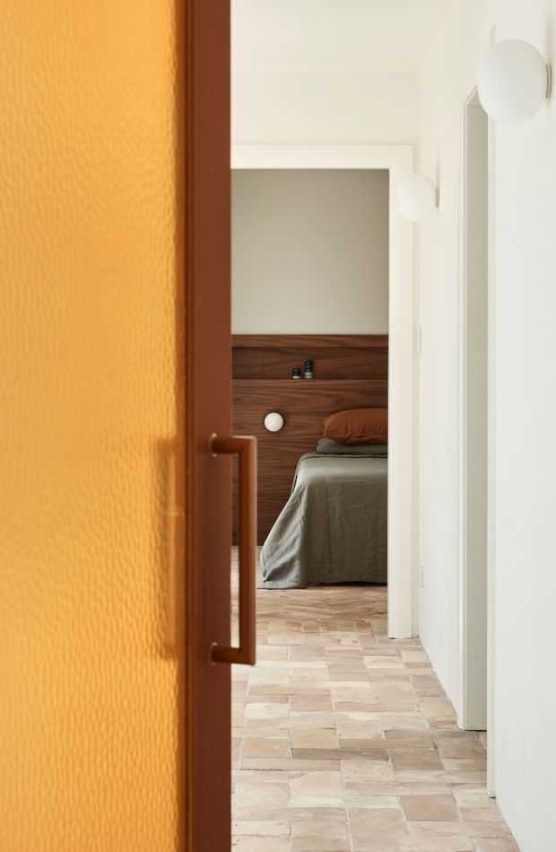 View through a partially open warm amber textured glass door into a minimalist bedroom. The narrow white hallway has spherical wall sconces, while the bedroom features a dark timber headboard wall, low bed with muted grey-brown bedding, and a pale irregular tiled or brick-like floor.