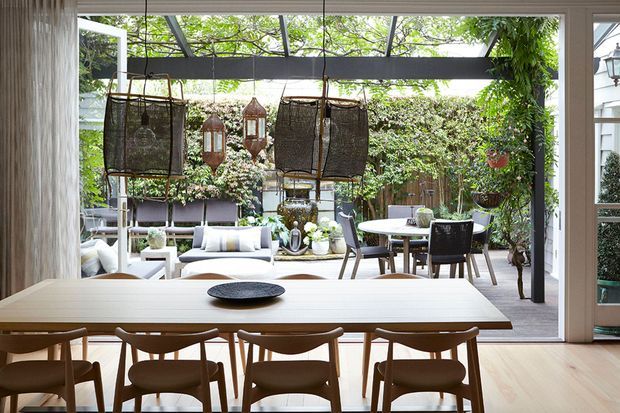 Bright open-plan dining area opening directly onto a lush landscaped courtyard through expansive floor-to-ceiling glazing. A long light timber dining table with matching timber chairs occupies the foreground, while an outdoor lounge and dining setting sits beyond on a paved terrace beneath a dark-framed pergola. Large woven pendant lights, abundant greenery, and neutral furnishings create a relaxed indoor-outdoor atmosphere.
