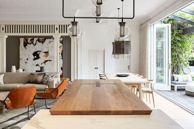 Bright open-plan dining and living area with a large solid timber dining table in the foreground, sculptural pendant lighting, white timber-lined ceiling and walls, built-in cabinetry, and a neutral sofa with orange leather lounge chairs. Full-height glazed doors open to a leafy outdoor terrace, creating a strong indoor-outdoor connection.
