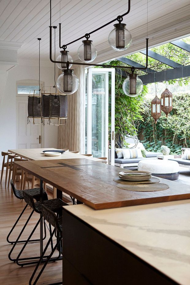A bright open-plan dining and kitchen interior with a long substantial timber dining table, black woven bar stools, a pale stone kitchen island, and distinctive glass globe pendant lighting with dark metal frames. White painted wall and ceiling panelling, timber flooring, and a large glazed opening connect the room to a leafy outdoor lounge. The space combines warm natural materials with crisp white surfaces and black accents.