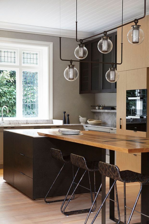 A refined kitchen interior featuring a large island with a warm timber benchtop and dark cabinetry, three bar stools, integrated black appliances, a stone-look splashback, and a bank of black-framed upper cabinetry. A decorative multi-light pendant with clear globes hangs above the island. The room has a white-lined ceiling, light timber flooring, neutral walls, and a leaded-glass window with garden views.