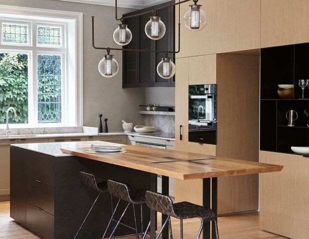A refined contemporary kitchen with a large timber island and integrated seating, dark cabinetry, pale wood wall joinery, stone benchtops, and a built-in oven. A decorative cluster of glass pendant lights hangs above the island, while tall leaded-glass windows bring in natural light.