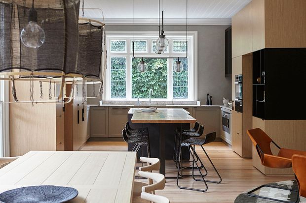 A contemporary kitchen and dining area with warm timber cabinetry, a central island with bar seating, a large multi-pane window, pendant lighting, and an adjacent dining table. The space combines pale timber floors and joinery with dark accents, black metal-framed stools, and sculptural dining chairs.