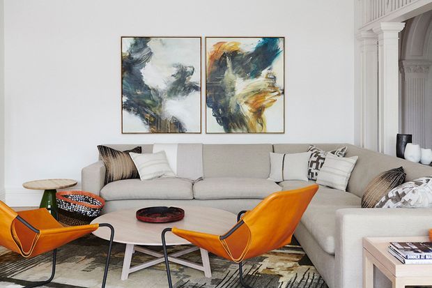 Bright, refined living room featuring a large light-grey sectional sofa, sculptural orange leather lounge chairs, a round pale timber coffee table, patterned rug, monochrome cushions, and abstract artwork. White architectural wall panelling and classical column details add character to the otherwise contemporary seating arrangement.