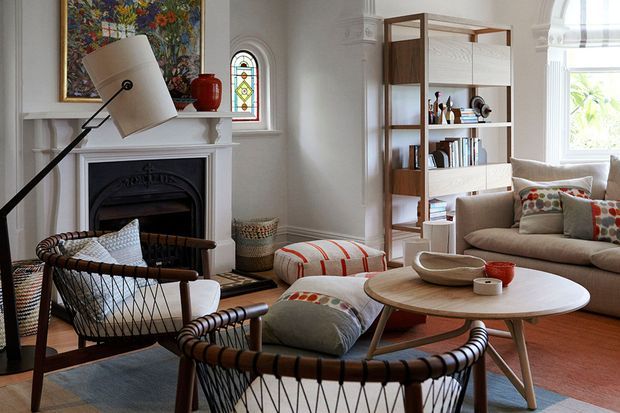 A warmly styled living room in a traditional home, featuring a decorative fireplace with white mantel, stained-glass windows, timber flooring, a freestanding timber bookshelf, and a mid-century-inspired seating arrangement with woven lounge chairs, a beige sofa, round timber coffee table, and patterned cushions.