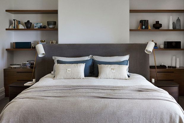 A refined contemporary bedroom centred on a neatly made bed with layered white and grey bedding, a dark upholstered headboard, and integrated bedside tables. Built-in timber shelving and cabinetry flank the bed, displaying books and decorative objects, with paired wall-mounted reading lights. The palette is restrained and neutral, creating a calm, polished atmosphere.