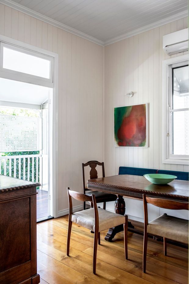 Bright dining area in a character home, featuring a traditional dark timber dining table and chairs, a built-in blue upholstered bench, warm timber floorboards, pale vertical wall panelling, and a white beadboard ceiling. The room opens onto a covered balcony through glazed doors and includes white-framed windows and a wall-mounted split-system air conditioner.