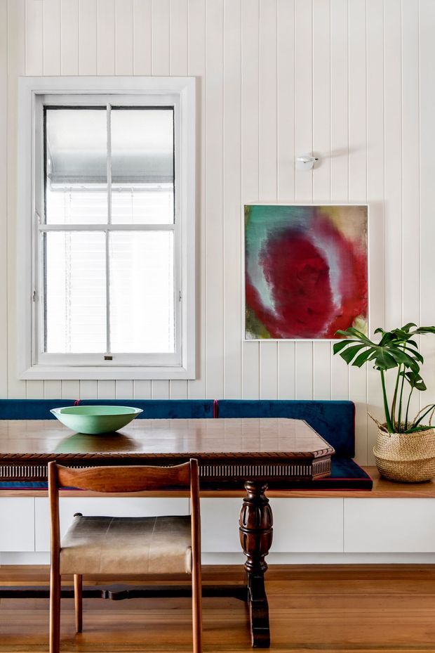 A bright, refined dining nook with a built-in deep-blue banquette beneath a framed abstract artwork. A timber dining table with turned legs sits in front of a vintage-style timber chair, while a white-framed window, vertical white wall panelling, timber flooring, and a potted indoor plant complete the scene.