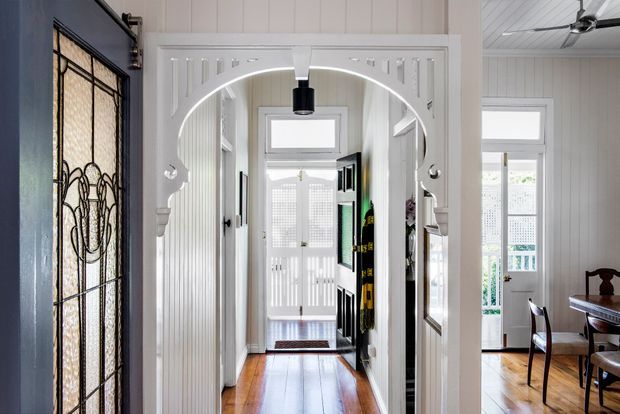 Bright traditional-style residential entry hall with a decorative white archway, white vertical wall panelling, polished timber floorboards, and a stained-glass front door at left. The hallway leads toward a glazed rear door and connects to a dining area on the right, with a ceiling fan and timber furniture visible.