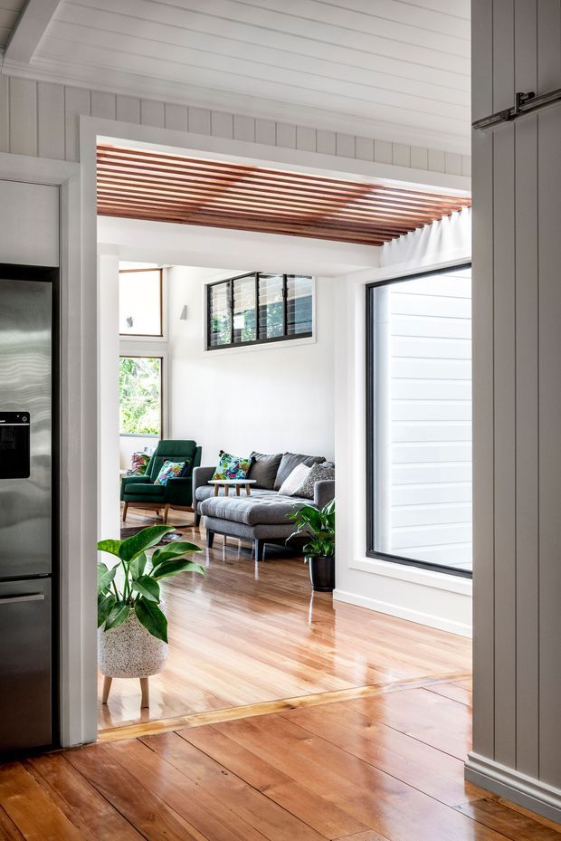 Bright interior view from a kitchen or adjoining circulation space into a living room. The room features polished timber floors, white vertical wall panelling and ceiling boards, a striking exposed timber slat ceiling detail, large black-framed windows, and a grey sofa with a green armchair. Indoor plants add a natural accent, while stainless-steel kitchen cabinetry is visible at the left edge.