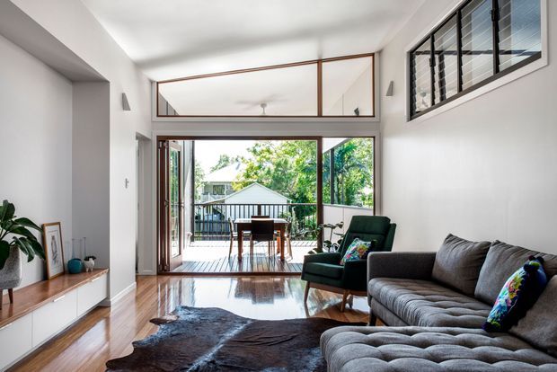 Bright, open-plan living room with polished timber flooring, a large grey sectional sofa, dark green armchair, low timber-and-white media cabinetry, and a cowhide rug. Full-height glazed doors open to a timber deck, while high clerestory glazing and a mirrored transom bring in abundant daylight. The room has a clean contemporary feel with strong indoor–outdoor connection.