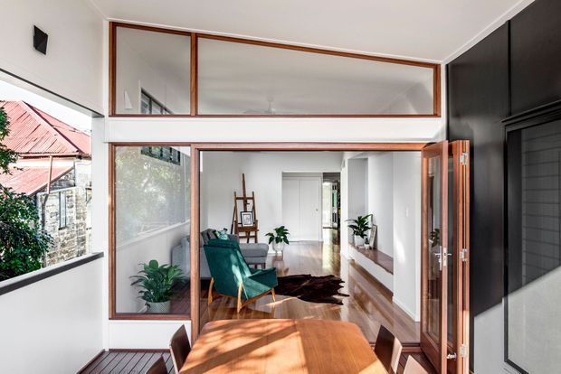 A bright contemporary interior viewed from a dining area, looking through timber-framed glazed partitions and an open door into a living room and hallway. The space features polished timber flooring, white walls, dark built-in wall panelling, a timber dining table, a teal lounge chair, indoor plants, and strong natural light. A red-roofed neighbouring building is visible through the glazing.