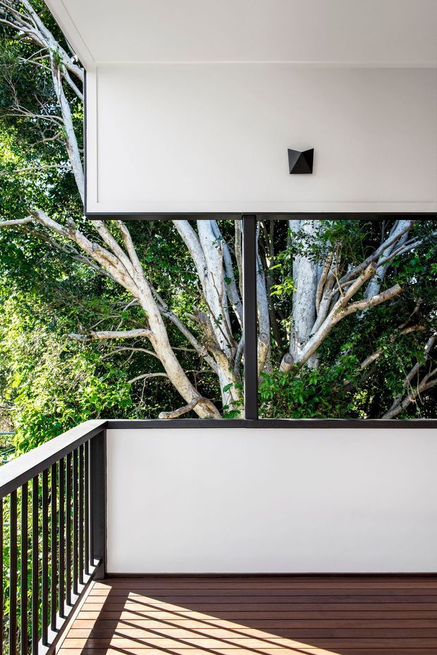 A bright, modern outdoor balcony or terrace surrounded by dense mature trees. The space features dark timber decking, black metal balustrades and posts, white rendered or painted wall and ceiling surfaces, and a minimalist black geometric wall light. Strong sunlight creates sharp shadows across the deck, with lush greenery visible beyond the railing.