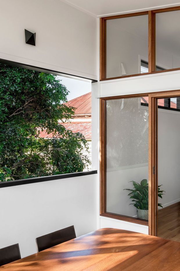 Bright interior view of a contemporary dining area opening toward a tall internal glazed void or courtyard edge. A large solid timber dining table dominates the foreground, with dark dining chairs, white walls, black-edged openings, and warm timber-framed glazing. A leafy tree and neighbouring terracotta roof are visible through the opening, while a potted plant and timber flooring add natural warmth. The space is light-filled and architecturally focused, with strong vertical glazing and open-plan connectivity.