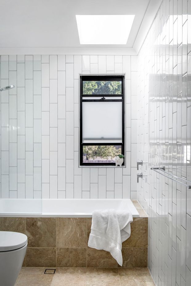 Bright, compact bathroom with a white bathtub, wall-mounted shower, toilet, chrome tapware and heated towel rail. White vertically arranged rectangular wall tiles cover the room, while the bathtub surround uses warm brown natural-stone-look tiles. A black-framed frosted window and large skylight provide natural light, with a small plant on the sill.