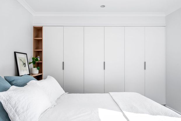 Bright, compact bedroom with a white bed and bedding in the foreground, a full wall of built-in white wardrobes with slim grey handles, and an integrated open timber shelving niche displaying a framed artwork and potted plant. The room has white walls, decorative ceiling trim, and a recessed downlight, creating a clean, calm contemporary look.
