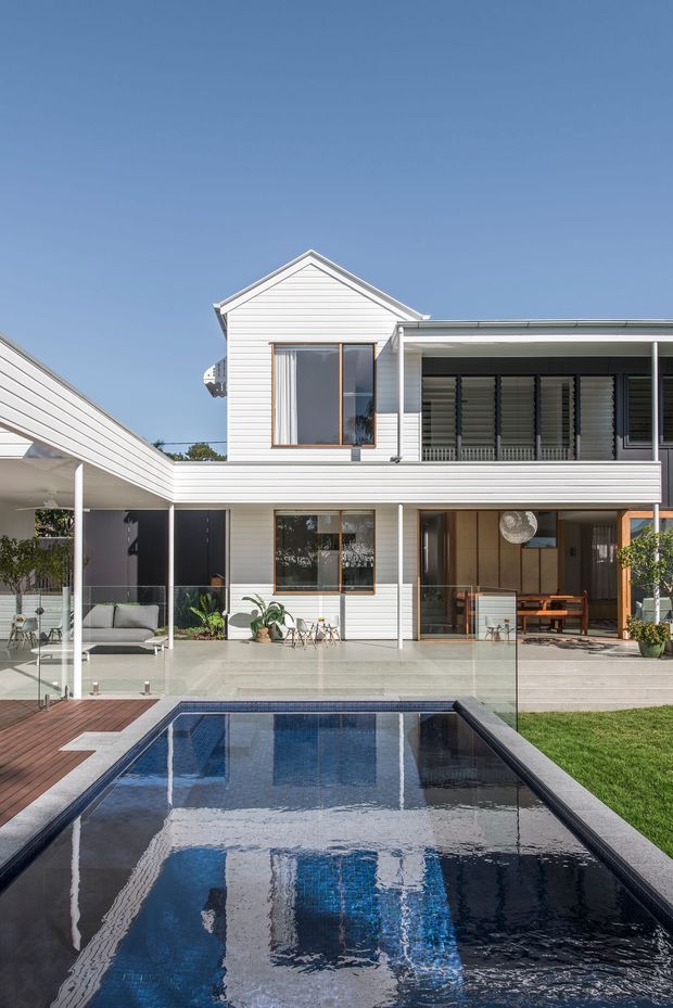 A bright contemporary multi-level residence viewed from the backyard, with white horizontal cladding, extensive glazing, covered outdoor living areas, a timber deck, and a rectangular swimming pool in the foreground. The home includes a gabled upper volume alongside a broad flat-roofed terrace structure, with landscaped planting and lawn around the pool.