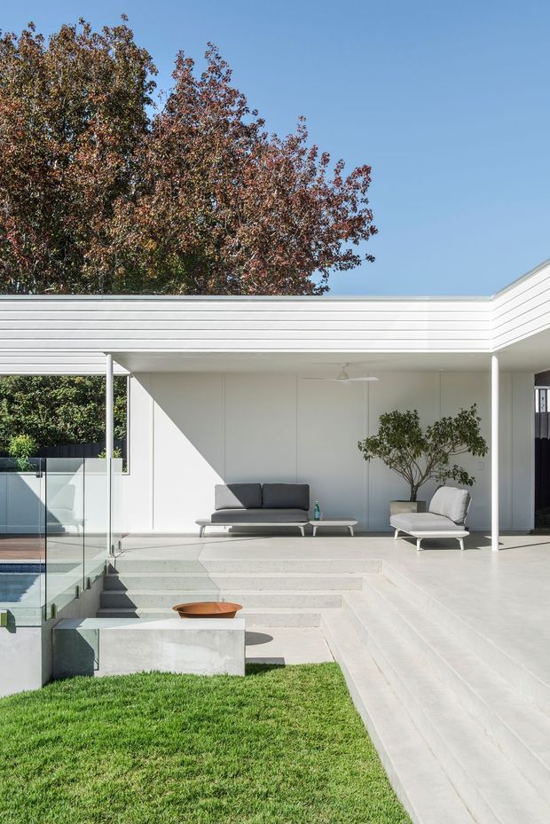 A bright, minimalist modern outdoor living terrace beside a swimming pool. The white flat-roofed structure has horizontal cladding, slender white columns, full-height glazing, broad pale steps and a covered lounge area furnished with a dark sofa, lounge chair and low table. A built-in concrete fire bowl feature sits beside the lawn, with mature trees visible beyond.