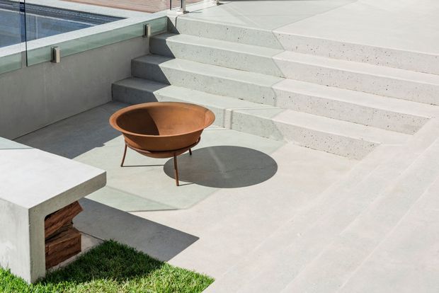 A sunlit contemporary outdoor terrace beside a swimming pool, featuring broad pale concrete steps, a glass pool balustrade, pale concrete paving, and a rust-coloured low fire bowl on slender legs. A minimalist concrete bench with timber storage or cladding sits at the edge of a small lawn area.