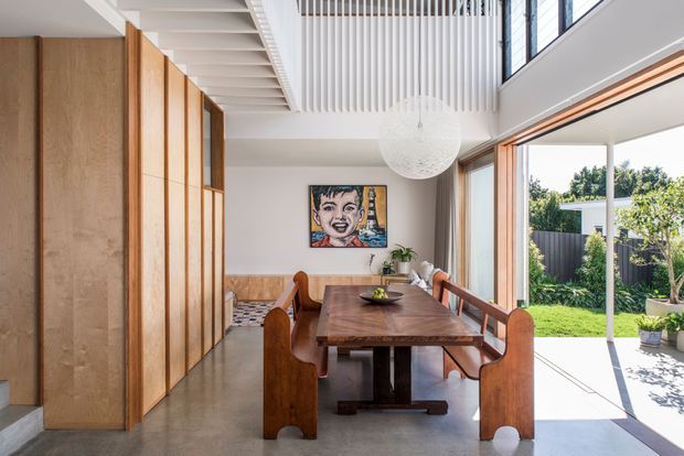 Bright contemporary dining area with a long solid-wood dining table and sculptural wooden seating, beside full-height glazing opening to a landscaped garden. The room features extensive timber built-in cabinetry or wall panelling, a polished concrete floor, a high ceiling with exposed white rafters and vertical slatted detailing, and a large textured spherical pendant light.