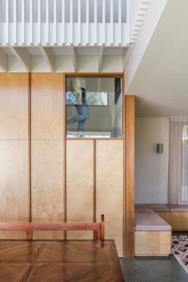 Interior view of a warm, highly detailed timber-lined space, likely focused on a dining area and built-in partition or storage wall. The foreground contains a dark timber dining table and matching bench, while the central wall features light plywood-style panels, darker timber framing, built-in cupboards, and a glazed internal opening reflecting the room. Above, a sculptural white ceiling or stair structure with repeated projecting slats and exposed beams creates strong geometric rhythm. A sitting area with built-in upholstered bench seating and curtains is visible to the right. The overall design is refined, tactile, and contemporary with strong custom joinery.