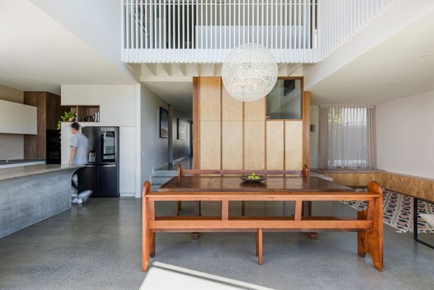 Open-plan contemporary interior combining a kitchen, dining area and living space. A large timber dining table and matching bench dominate the foreground, with a dark stone kitchen island and integrated cabinetry to the left. A timber-framed glazed partition and vertical white balustrade define a double-height void above, complemented by a large spherical pendant light and sheer curtains. The palette is light and neutral with warm timber accents and polished grey flooring.