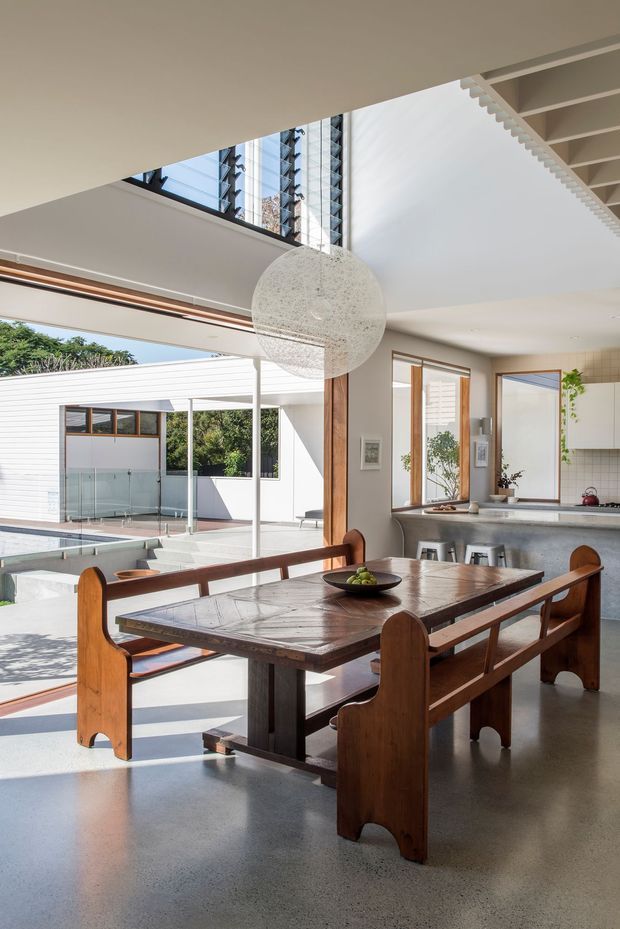 A bright, open-plan dining area with a substantial timber dining table and matching bench seating, adjoining a contemporary kitchen. The room has polished concrete flooring, white walls and cabinetry, warm timber window and door frames, and a large spherical pendant light. Extensive glazing and open sliding doors connect the interior to a covered outdoor courtyard and swimming pool, while a dramatic high-level opening and exposed stair structure add architectural interest.