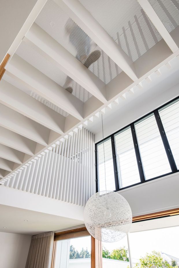 Bright contemporary interior looking upward toward a dramatic open stair or mezzanine structure. White exposed beams and perforated panels create a patterned overhead ceiling, with vertical white balustrade slats, high clerestory-style windows, timber-framed glazing, and a large spherical pendant light. The space appears airy and double-height with strong natural light and a predominantly white palette accented by timber and dark window frames.