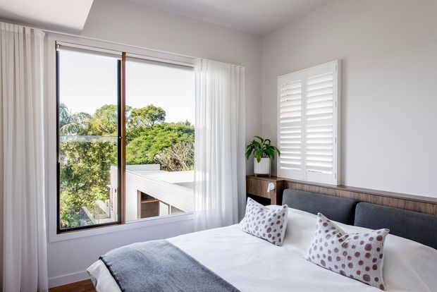 Bright, contemporary bedroom with a neatly made bed, grey upholstered headboard, white bedding, patterned cushions and a grey throw. White walls, sheer curtains, plantation shutters and a timber-framed window provide a clean, light-filled setting with leafy views outside. A slim timber bedside ledge and potted plant add warmth.