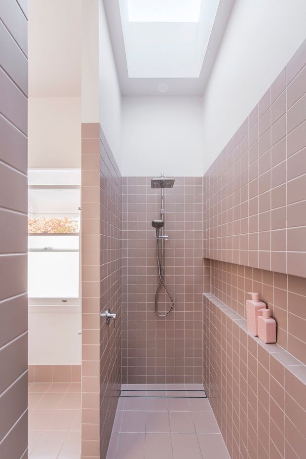 A narrow contemporary walk-in shower bathroom clad in small dusty pink square tiles from floor to ceiling. A rainfall shower and handheld shower are mounted on the rear tiled wall, with a tiled built-in bench and recessed shelf holding pink bottles on the right. A large rectangular skylight brings in abundant natural light, and a partially visible white vanity and window appear beyond the shower enclosure.