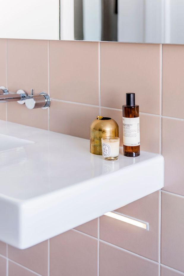 Close-up of a compact bathroom washbasin with a glossy white integrated basin and wall-mounted chrome tapware. The walls are clad in pale blush-pink square tiles with light grout, and a mirrored cabinet or mirror is visible above. Amber and gold toiletry bottles sit on the basin ledge, with a slim horizontal wall light or illuminated recess below.
