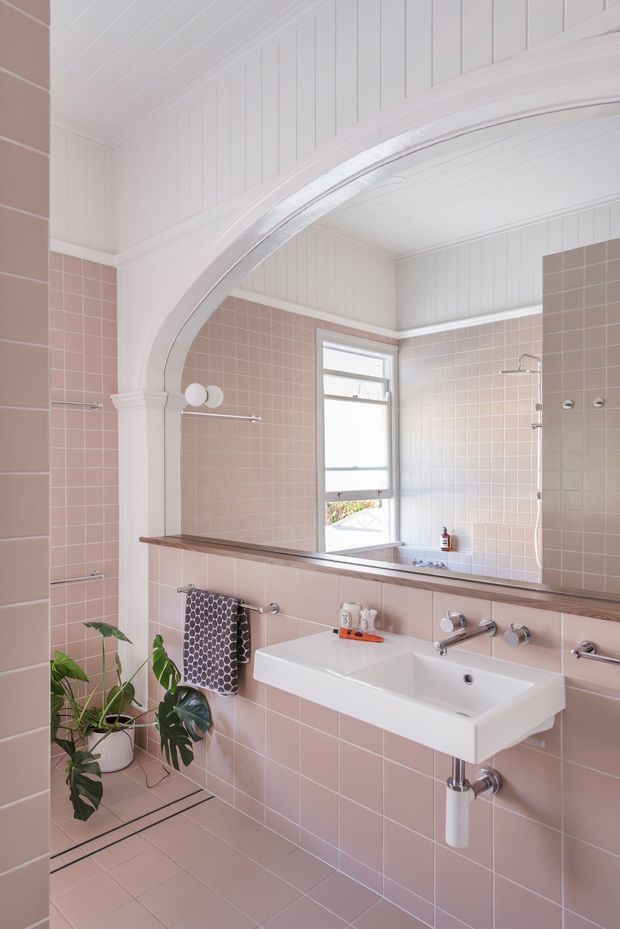 A bright, refined bathroom with blush-pink tiled walls and floor, white vertical wall panelling and a white panelled ceiling. A large curved arch frames the vanity and shower area, with a white wall-mounted basin, timber-toned countertop ledge, chrome tapware, frameless shower glazing, and a tall window. A potted monstera and patterned towels add softness and contrast.